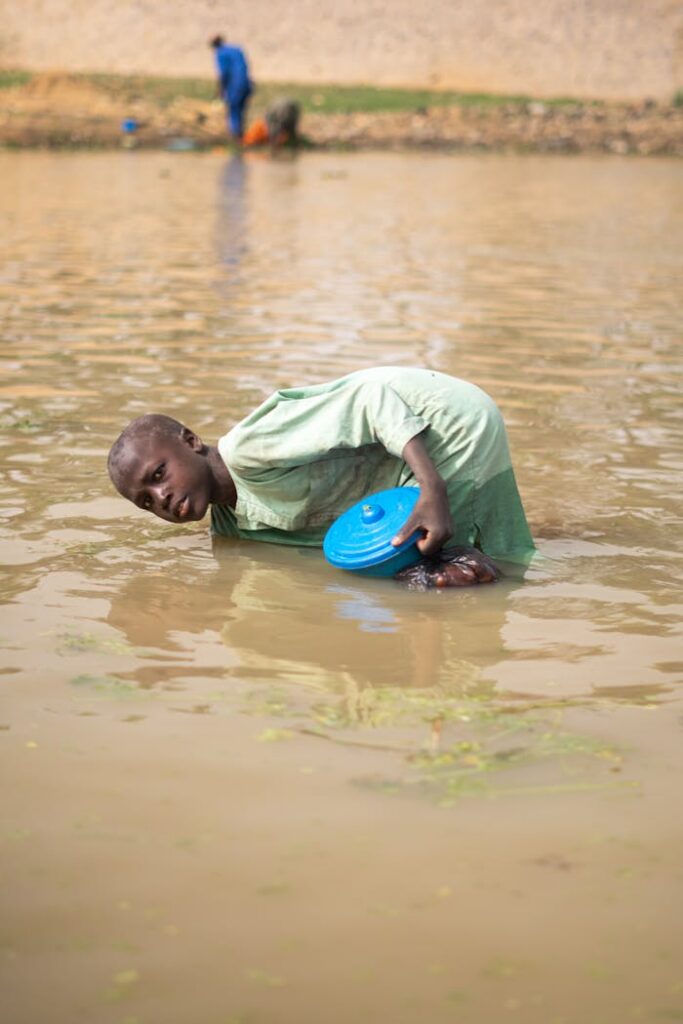 Young boy wading in a flood with a blue bowl collecting water. Outdoors scene.