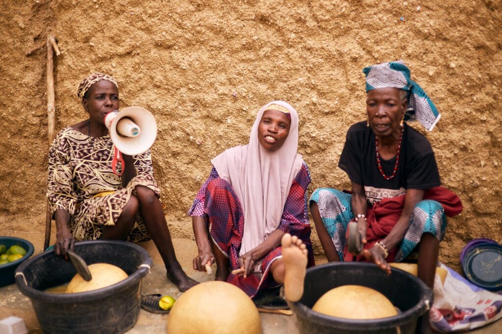 Three African women sitting outdoors preparing traditional food using local tools.